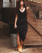 Woman in a black dress and cowboy hat standing in a rustic setting with wooden walls and hay.